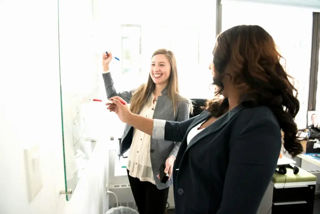 Two professional women discussing ideas on a whiteboard in a modern office setting.