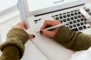 From above crop unrecognizable female in casual warm sweater taking notes in opened notebook with stylish silver pen while using modern netbook